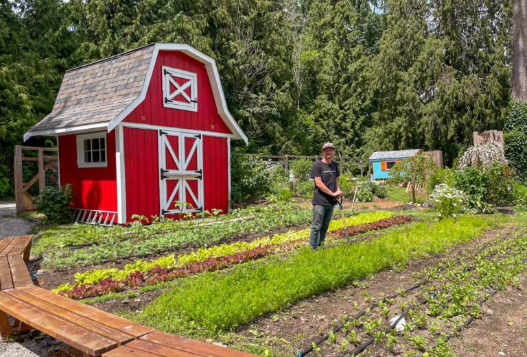 A man stands in the One Tiny Farm garden, working with garden tools, in front of a red barn.