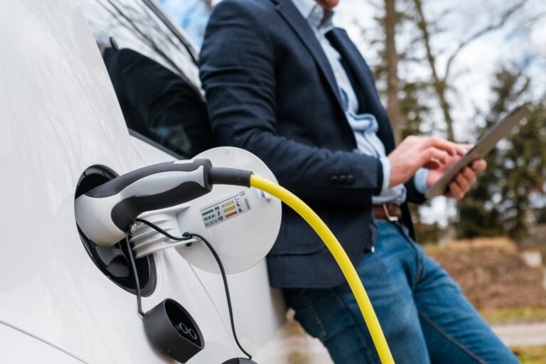 A stock image of an electric car charging with a person waiting in the background working on an ipad.